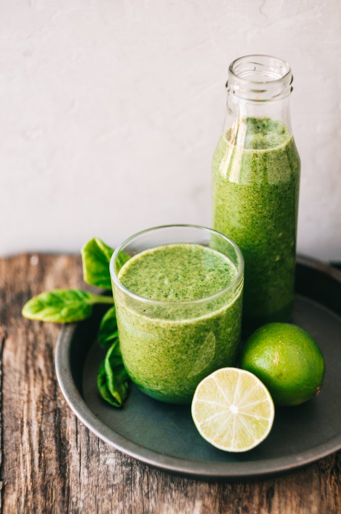 Un jus vert sur un plateau accompagné de feuilles d'épinards et de citrons verts